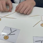 Mâinile unui copil care reproduce un model cu bețișoare, după cartonașe așezate pe masă
